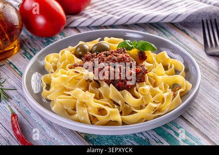 Piatto di tagliatelle condito con ricco sugo di carne, olive verdi e basilico. Servito su un tavolo rustico di legno con pomodori e peperoncino. Primo piano Foto Stock