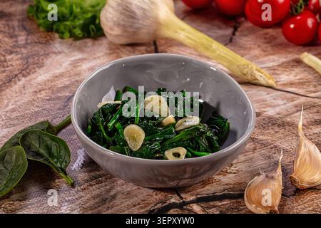 Spinaci verdi freschi saltati con spicchi d'aglio tagliati in una ciotola grigia, circondati da aglio crudo e pomodori su una superficie di legno, salutare vegetariano Foto Stock