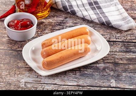 Tre salsicce di frankfurter disposte su un piatto rettangolare bianco, accompagnate da una ciotola di ketchup e una bottiglia di olio, poste su un rustico tavolo di legno W. Foto Stock