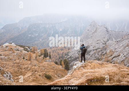 Escursionista che si affaccia su aspri terreni calcarei e foreste autunnali nelle Dolomiti. Paesaggio montano con scogliere rocciose, larici dorati e cime nebbiose Foto Stock
