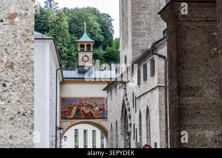 Una vista attraverso un arco nella città vecchia di Salisburgo verso una storica torre dell'orologio con una cupola di rame e una faccia dorata dell'orologio, nonché un affresco sulla facciata di un edificio; sulla destra, l'imponente parete esterna della Chiesa francescana. La città vecchia di Salisburgo è un sito patrimonio dell'umanità dell'UNESCO dal 1997. Salisburgo, Austria Foto Stock