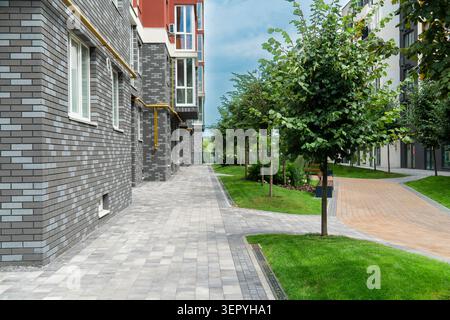 Accogliente cortile di edifici residenziali moderni con facciata in mattoni grigi, prati verdi, giovani alberi e percorsi pedonali lastrati in una giornata estiva Foto Stock
