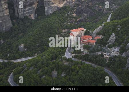 Vista aerea del Monastero di Rousanou a Meteora, arroccato su una ripida colonna rocciosa circondata da lussureggianti foreste verdi e da una tortuosa strada di montagna a Tri Foto Stock