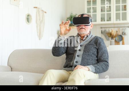 Un uomo anziano entusiasta indossando occhiali VR in realtà virtuale metaverse a casa. Nonno che tocca l'aria durante l'esperienza di realtà virtuale su casco per realtà virtuale. Videogame di simulazione ad alta tecnologia Foto Stock
