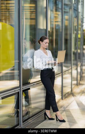Una donna si trova accanto a una grande parete di vetro di un edificio moderno. Sta usando un notebook, vestito con abiti da lavoro. La luce del sole è presente, evidenziando il suo testamento Foto Stock