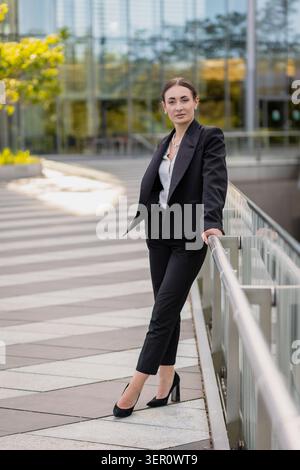Una donna si trova accanto a una ringhiera che indossa un abito nero di fronte ad un edificio moderno. Posa con sicurezza mentre guarda la telecamera, con gli alberi nella B. Foto Stock