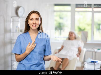 Medico anziano sorridente in uniforme con paziente donna che giace dietro in clinica. Foto Stock