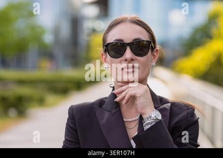 Una donna sta fuori sotto il sole con gli occhiali da sole. Ha i capelli lunghi ed è vestita con un abito scuro. Lo sfondo mostra piante verdi e moderne Foto Stock