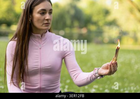 Una donna siede sull'erba in un parco con un bastone in fiamme. La luce del sole splende e gli alberi sono sullo sfondo mentre si concentra sulla fiamma. Foto Stock