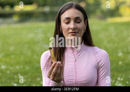 Una donna siede pacificamente in un parco con gli occhi chiusi. Tiene l'incenso in una mano, concentrandosi sulla sua meditazione sotto il cielo limpido in mezzo al verde gra Foto Stock