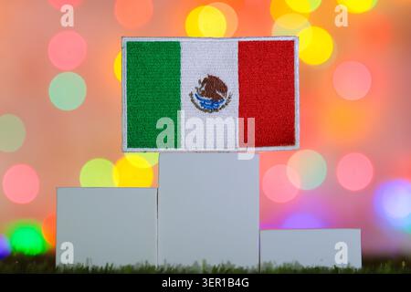 Bandiera nazionale del Messico su un podio bianco a blocchi disposto nella più alta posizione al primo posto su uno sfondo colorato di luci bokeh soffici. Foto Stock