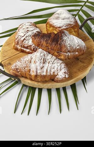 Croissant appena sfornati cosparsi di zucchero a velo, appoggiati su un vassoio di legno rustico adornato con foglie di palma, creando un ambiente tropicale per la colazione Foto Stock