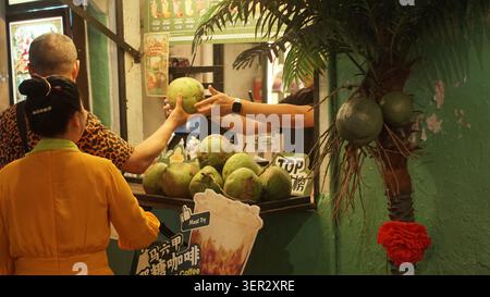 Fornitore che passa una noce di cocco fresca a un cliente in un mercato notturno a Malacca, Malesia, con bevande tropicali in vendita. Foto Stock