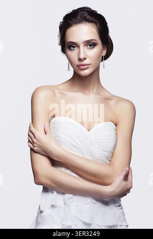 Ritratto di giovane e bella bruna donna sposa in bianco abito da sposa con mani incrociate su sfondo grigio chiaro Foto Stock