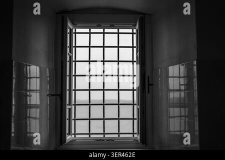 Finestra interna aperta con barre di sicurezza in metallo e luce riflessa sulle pareti piastrellate. Dettagli architettonici con simmetria, contrasto e lin geometrico Foto Stock