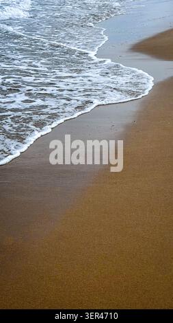 Le dolci onde oceaniche si infrangono su una spiaggia sabbiosa, creando una costa liscia con sabbia bagnata e acqua spumosa, catturate in un ambiente costiero sereno Foto Stock