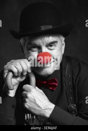 Ritratto a colori selettivo di clown ridendo appoggiato sul manico di un ombrello. Un uomo anziano con naso rosso da clown e cappello da bowler sulla testa. Vestito Foto Stock