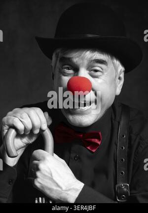 Ritratto a colori selettivo di clown ridendo appoggiato sul manico di un ombrello. Un uomo anziano con naso rosso da clown e cappello da bowler sulla testa. Vestito Foto Stock
