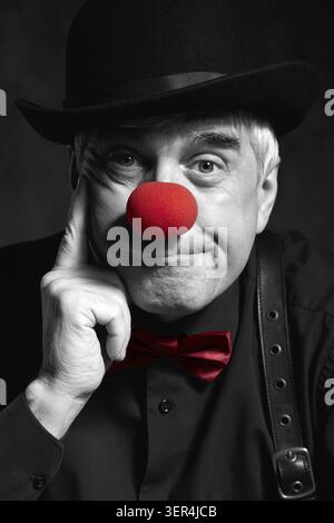 Spettacolare ritratto a colori selettivo di un pagliaccio triste su sfondo scuro. Un uomo anziano con naso rosso da clown e cappello da bowler sulla testa. Vestito con blac Foto Stock