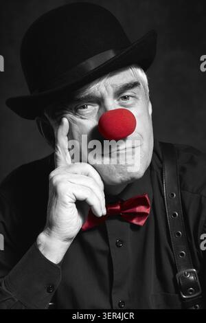 Spettacolare ritratto a colori selettivo di un pagliaccio triste su sfondo scuro. Un uomo anziano con naso rosso da clown e cappello da bowler sulla testa. Vestito con blac Foto Stock