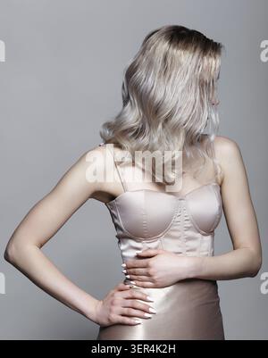 Ritratto di una giovane donna in abito beige con spalline. I peli stanno chiudendo il viso. Donna bionda con le mani in vita su sfondo grigio Foto Stock