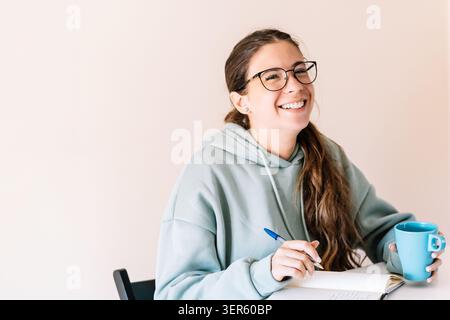 Una donna che indossa gli occhiali è seduta a un tavolo con una tazza blu e una penna. Lei sorride ed è felice Foto Stock