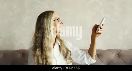 Felice sorridente donna matura caucasica accende il condizionatore d'aria utilizzando il telecomando nel soggiorno di casa Foto Stock