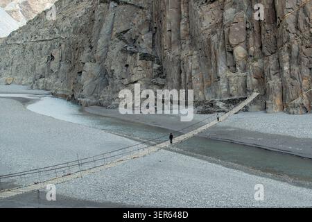 Vista del ponte sospeso Hussaini che attraversa il fiume Hunza con ripide scogliere rocciose e persone che camminano attraverso le tavole di legno di Hussaini, Gilgit, Paki Foto Stock