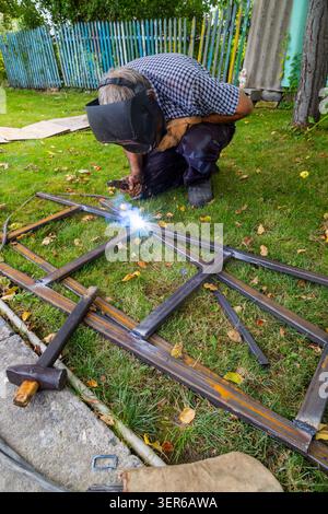 Uomo maturo si inginocchia all'aperto, saldando il telaio in metallo nel cortile posteriore. Le scintille volano mentre si concentra sulla lavorazione dei metalli, circondato da foglie autunnali. Foto Stock