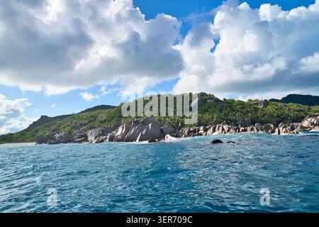 Panorama costiero mozzafiato di La Digue, Seychelles, da una barca, con iconici rocce di granito, colline verdi lussureggianti e acque oceaniche di un blu profondo sotto un cielo nuvoloso Foto Stock