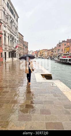 Donna che cammina con un ombrello accanto a un canale in una giornata di pioggia a Canareggio Venezia, Italia. Ha trent'anni e indossa un trench coat. Foto Stock