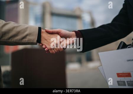 Stretta di mano tra due professionisti all'aperto con documenti vicino agli edifici della città Foto Stock