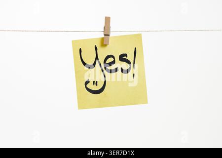 Scritto a mano "Sì!" Su una nota adesiva gialla ritagliata su una corda, con una giocosa faccia sorridente integrata nella lettera Y, isolata su una schiena bianca Foto Stock