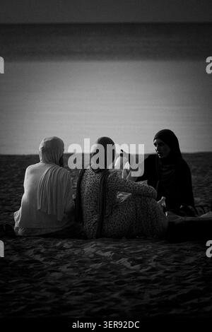 Tre donne musulmane in hijab tradizionali sedute su una spiaggia sabbiosa al tramonto, che parlano e si rilassano. Fotografia in bianco e nero. Foto Stock