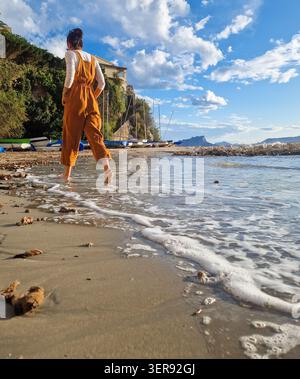 Vista posteriore di una donna che cammina a piedi nudi lungo una spiaggia mediterranea in estate, con onde dolci, litorale sabbioso, barche a vela e scogliere sotto un luminoso b Foto Stock