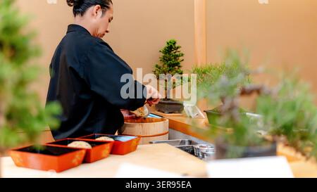 Abile sushi chef in uniforme tradizionale preparando nigiri freschi al banco di un ristorante giapponese a Tokyo, con vassoi bento, vasca di legno e bonsai Foto Stock