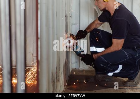 Un giovane uomo caucasico lavora con una sega circolare, tagliando attraverso il muro di un container di spedizione. Le scintille volano mentre costruisce una casa da contenimento marino Foto Stock