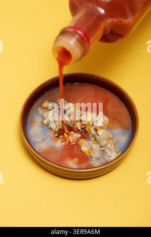 Primo piano di tapas spagnole: I cockles marinati in una piccola ciotola mentre la salsa viene versata da una bottiglia su uno sfondo giallo brillante, perfetto per l'aperitivo e. Foto Stock