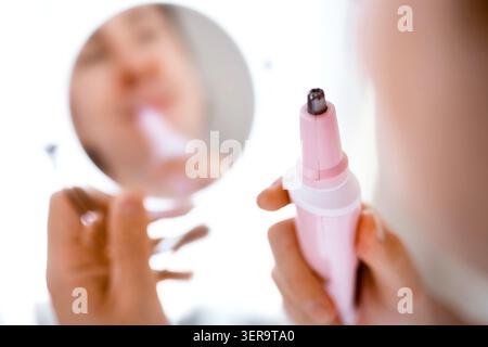 Primo piano di una mano maschile che tiene in mano un rifinitore elettrico per la rimozione dei peli del naso. Il riflesso a specchio mostra la cura del viso e la routine di cura del viso di un uomo. Alta qualita' Foto Stock