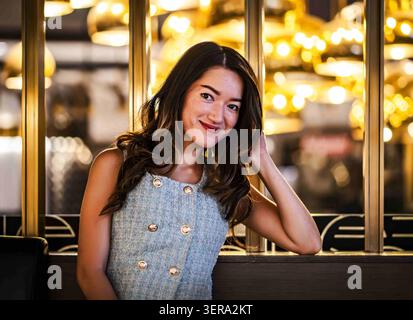 Giovane donna sorridente seduta in un ristorante elegante con calde luci accese, abiti eleganti e posa rilassata, creando un'accogliente vita notturna e uno stile di vita Foto Stock