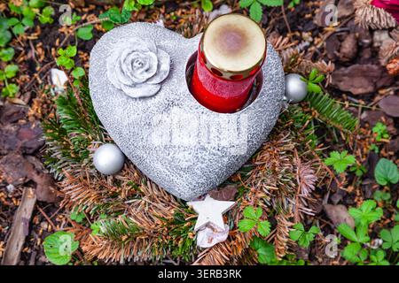 Porta tomba in pietra a forma di cuore con candela commemorativa rossa e rosa scolpita su un terreno con rami di conifere Foto Stock