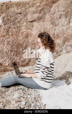 Spiaggia per laptop freelance. Una giovane donna lavora a distanza mentre è seduta su una spiaggia di ciottoli di fronte a un'enorme parete rocciosa Foto Stock
