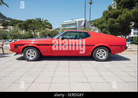 Vista a profilo laterale completa di una muscle car Ford Mustang Mach 1 rossa d'epoca con strisce nere da corsa parcheggiata su una passeggiata pavimentata in una giornata di sole. Foto Stock