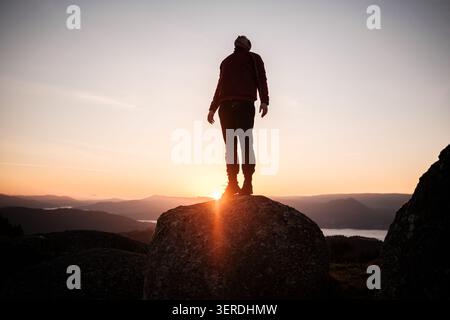 Uomo in piedi stagliato su una roccia, guardando il cielo del tramonto su un lontano lago di montagna Foto Stock