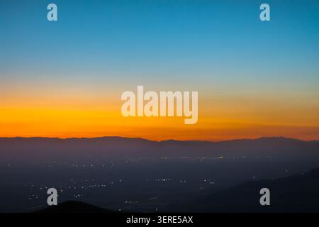 Il tranquillo paesaggio crepuscolare presenta un cielo vibrante che passa dal blu intenso all'arancione e al giallo. Le montagne sagomate incorniciano una lontana città di Bishkek Foto Stock
