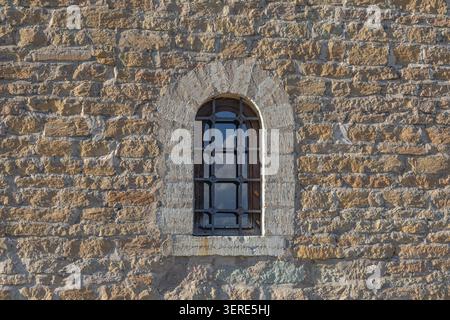 Dettaglio di una stretta finestra ad arco con barre di metallo incastonate in spesse mura della vecchia fortezza in pietra. Elemento architettonico storico Foto Stock
