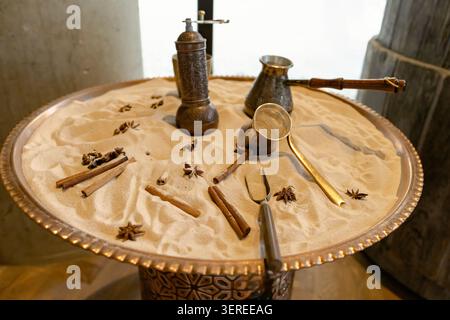 Set per la preparazione del caffè turco tradizionale con pentole in rame, macinacaffè manuale, cannella, anice stellato disposto su vassoio di sabbia caldo L'illuminazione calda mette in risalto le texture metalliche e le spezie aromatiche Foto Stock