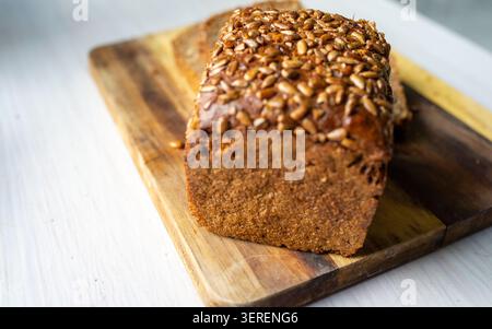 Pane rettangolare a grana multipla appena sfornato posto su un tagliere rustico in legno, crosta ricoperta di semi di girasole tostati e cereali interi, cru denso Foto Stock