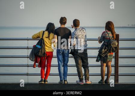 Un gruppo di quattro giovani individui è visto da dietro, appoggiato contro una ringhiera di metallo su un lungomare a Baku, Azerbaigian. Stanno guardando Foto Stock