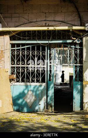 Una persona sagomata si trova all'interno di un corridoio poco illuminato, visto attraverso un cancello in ferro battuto turchese, parzialmente aperto, a Baku, Azerbaigian. Giallo Foto Stock
